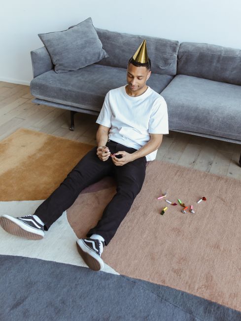 A young man wearing a white t-shirt, black pants, and sneakers is sitting on a multi-colored area rug in a living room, with a gold party hat on his head. He is holding a small object in his hands, an